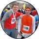 Two men in athletic wear smile at the camera after a race. The man on the left wears a red shirt and has race number 162, while the man on the right wears an orange shirt with race number 1159. Other participants are visible in the background.